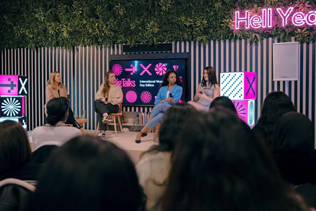 Four women are seated on a stage with a leafy backdrop and a neon sign. They are engaged in a panel discussion. The audience is visible in the foreground, and colorful decorative elements are on either side of the stage.